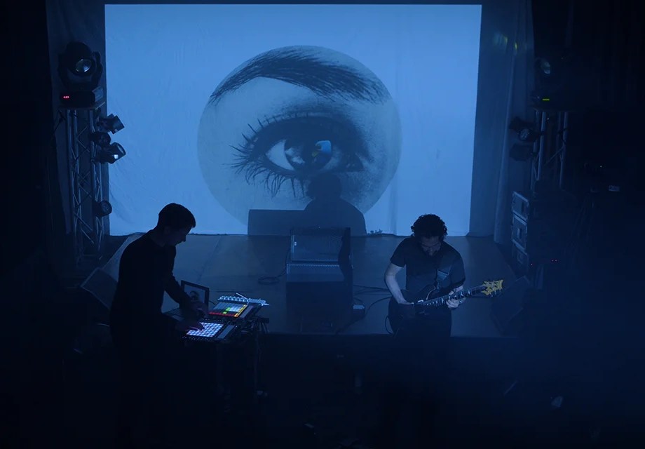 two people on stage, with visuals and a guitar