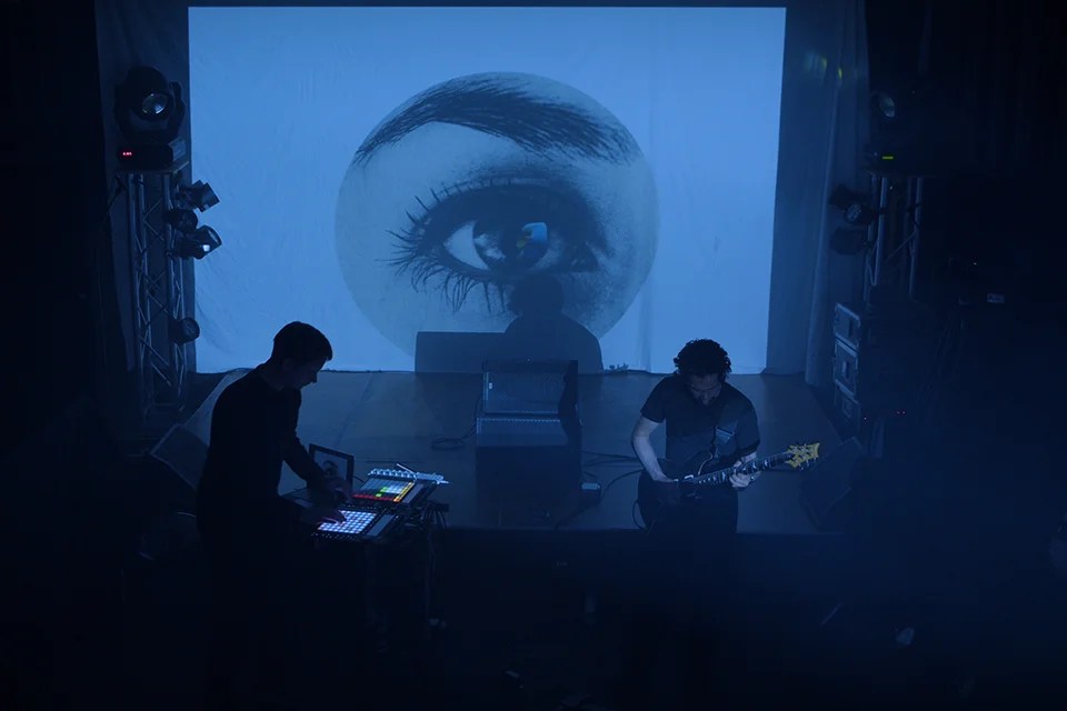 two people on stage, with visuals and a guitar
